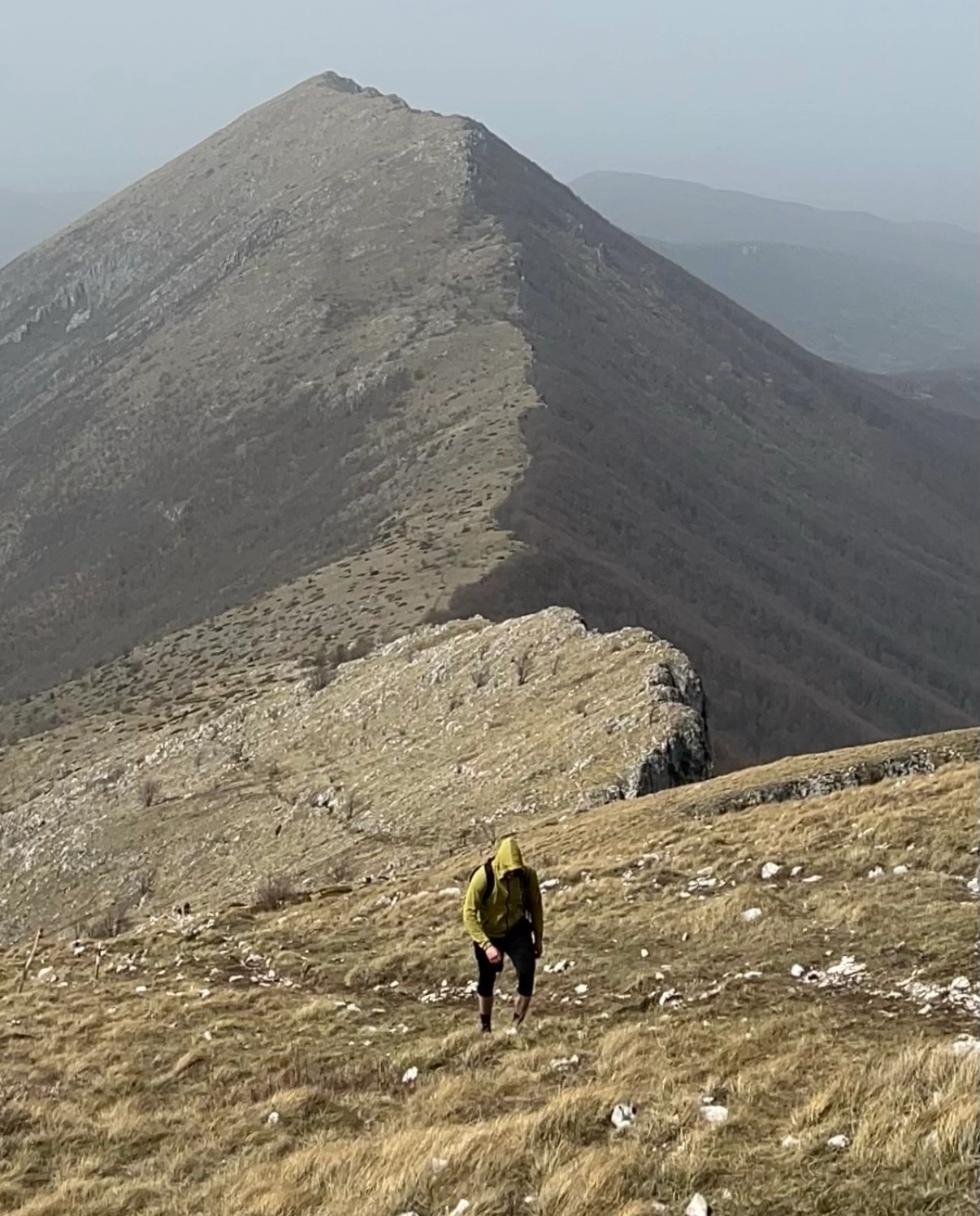 Person in a yellow hoodie hiking uphill on a grassy mountain ridge under a hazy sky.