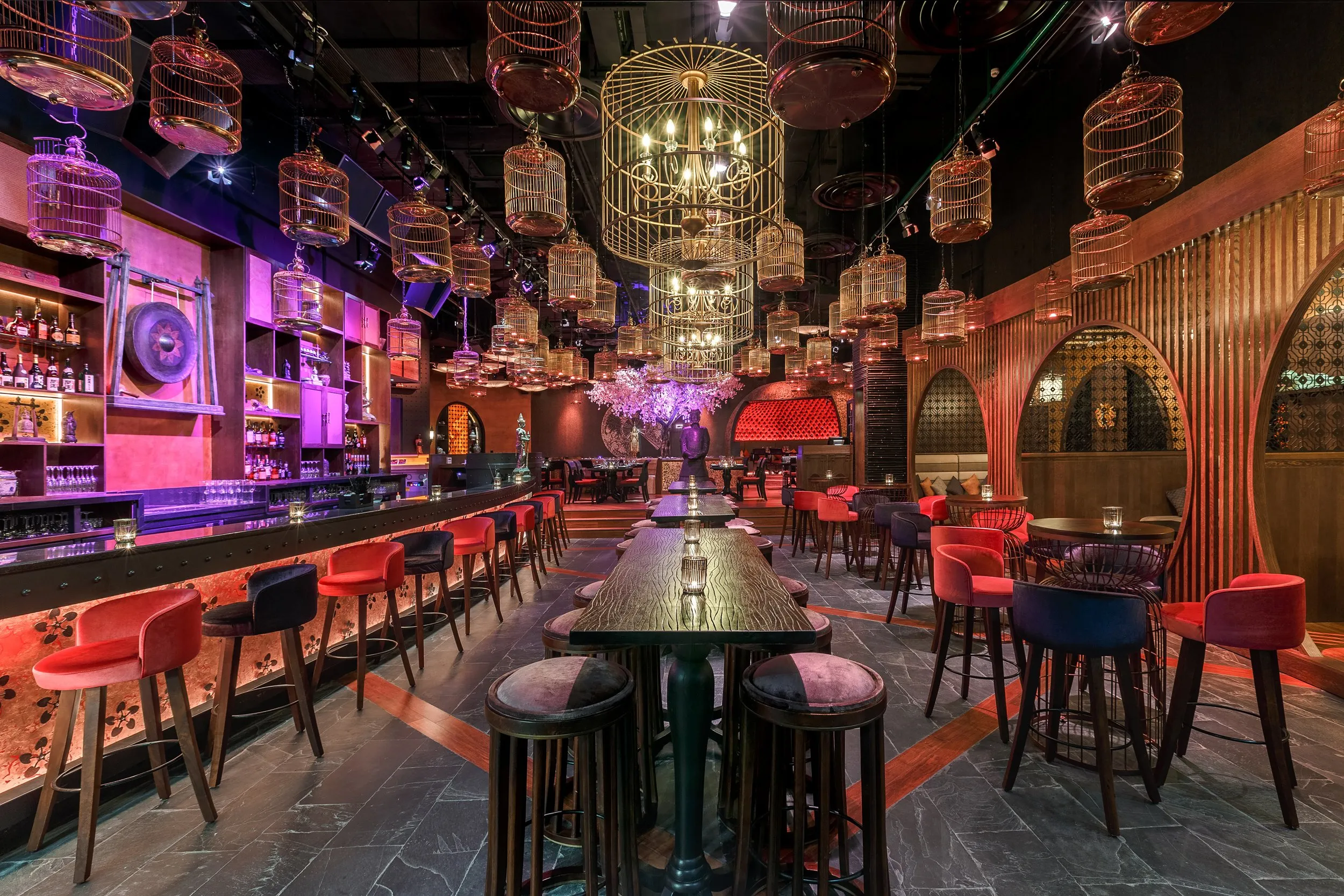 Stylish bar interior with red and black stools, wooden tables, birdcage-style chandeliers, and illuminated shelves with bottles.