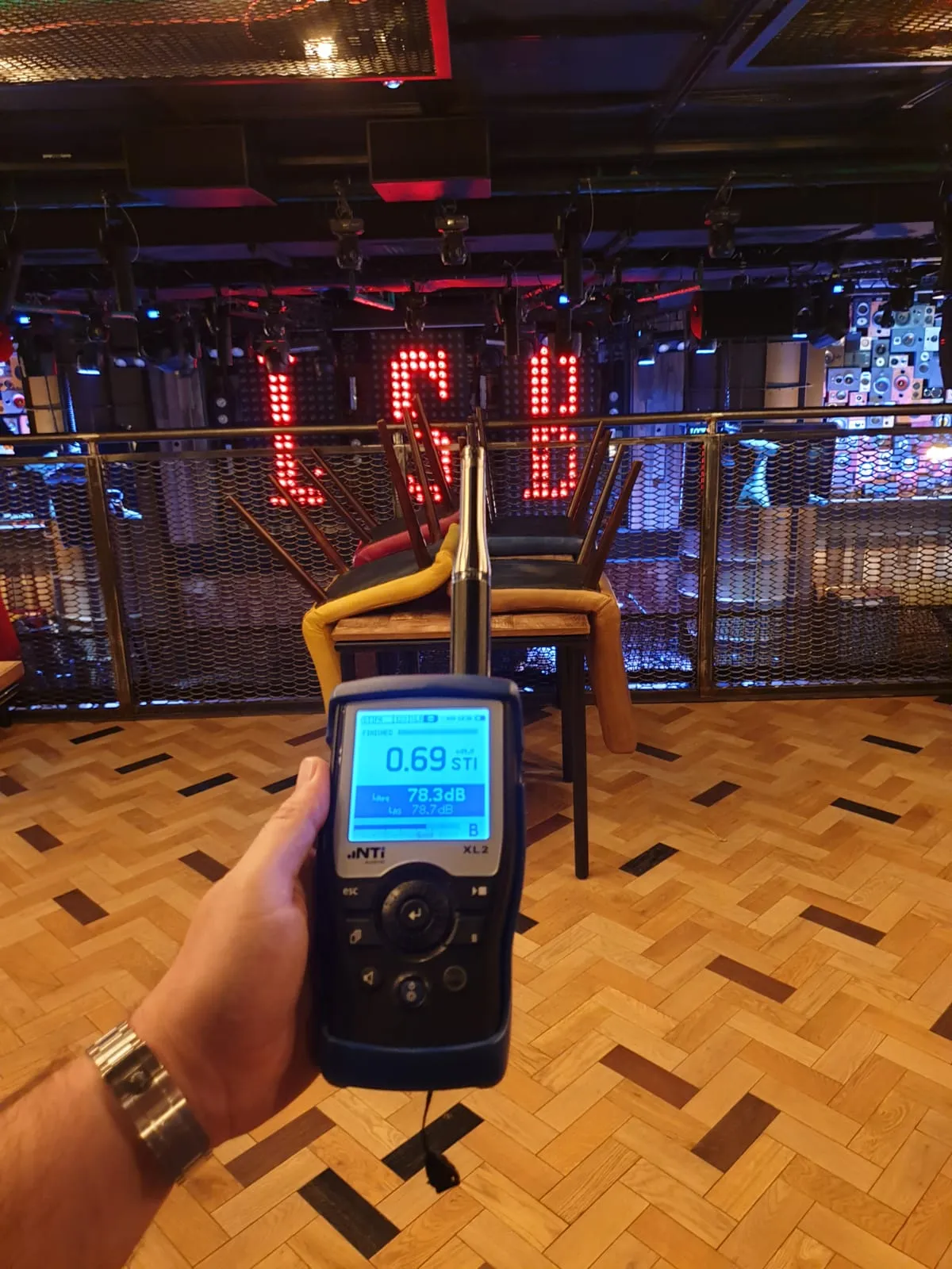 Hand holding a sound level meter showing 78.3 dB in a dimly lit room with chairs stacked on tables and illuminated letters in the background.