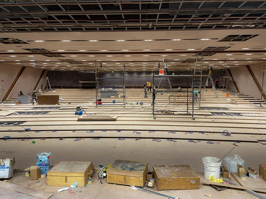 Large indoor auditorium under construction with wooden flooring being installed and scaffolding in the centre.
