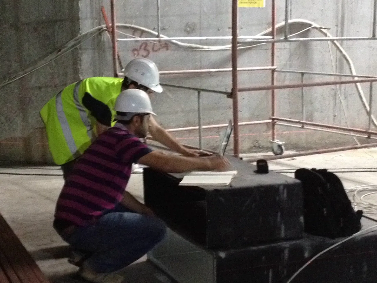 Two construction workers wearing white helmets, one in a yellow safety vest, working on a laptop and notebook inside a building under construction.