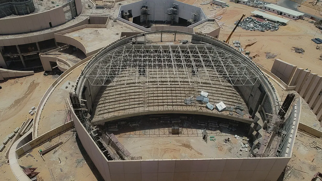 Aerial view of a large circular building under construction with a partially completed steel framework roof and tiered seating inside.