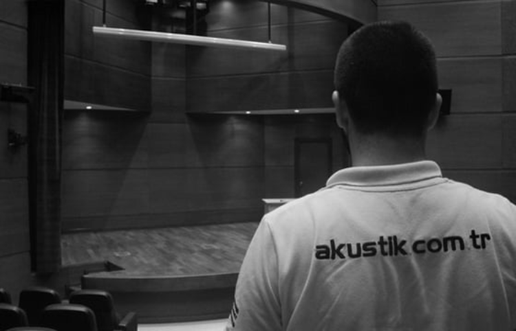 Person wearing a white shirt with 'akustik.com.tr' written on the back, facing an empty auditorium stage in a dimly lit room.