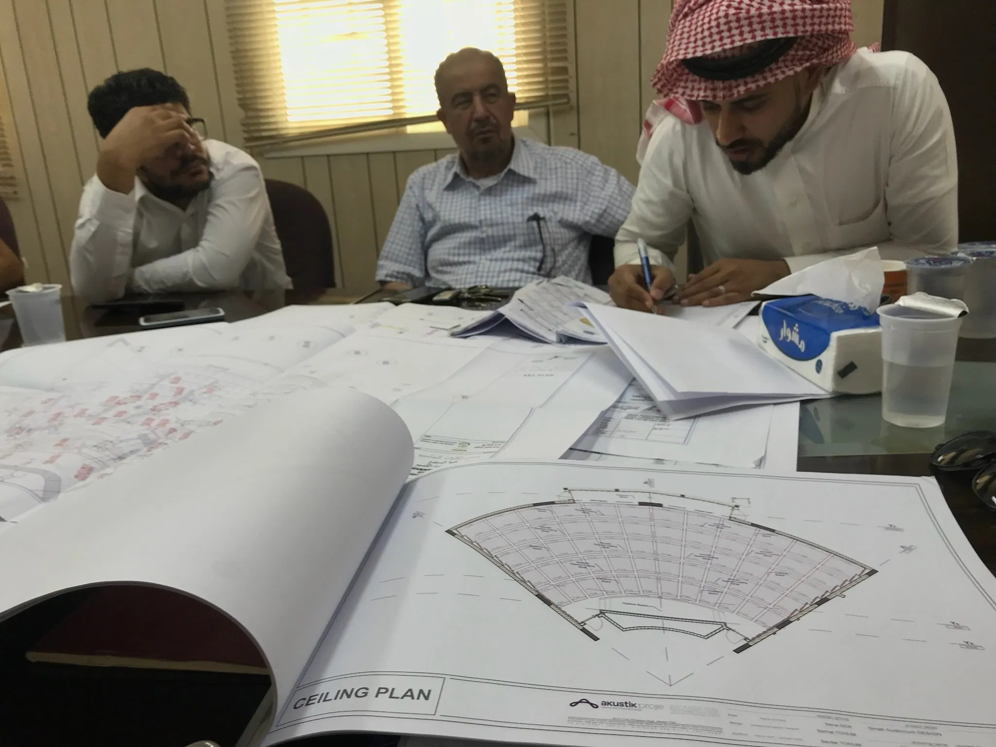 Three men reviewing architectural ceiling plans spread out on a table, with one man writing on a document.