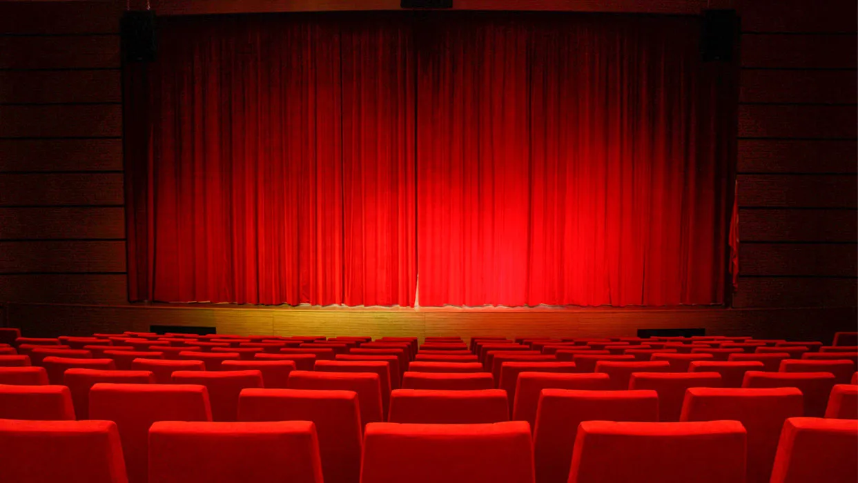 Empty theatre with rows of red seats facing a stage with closed red curtains.