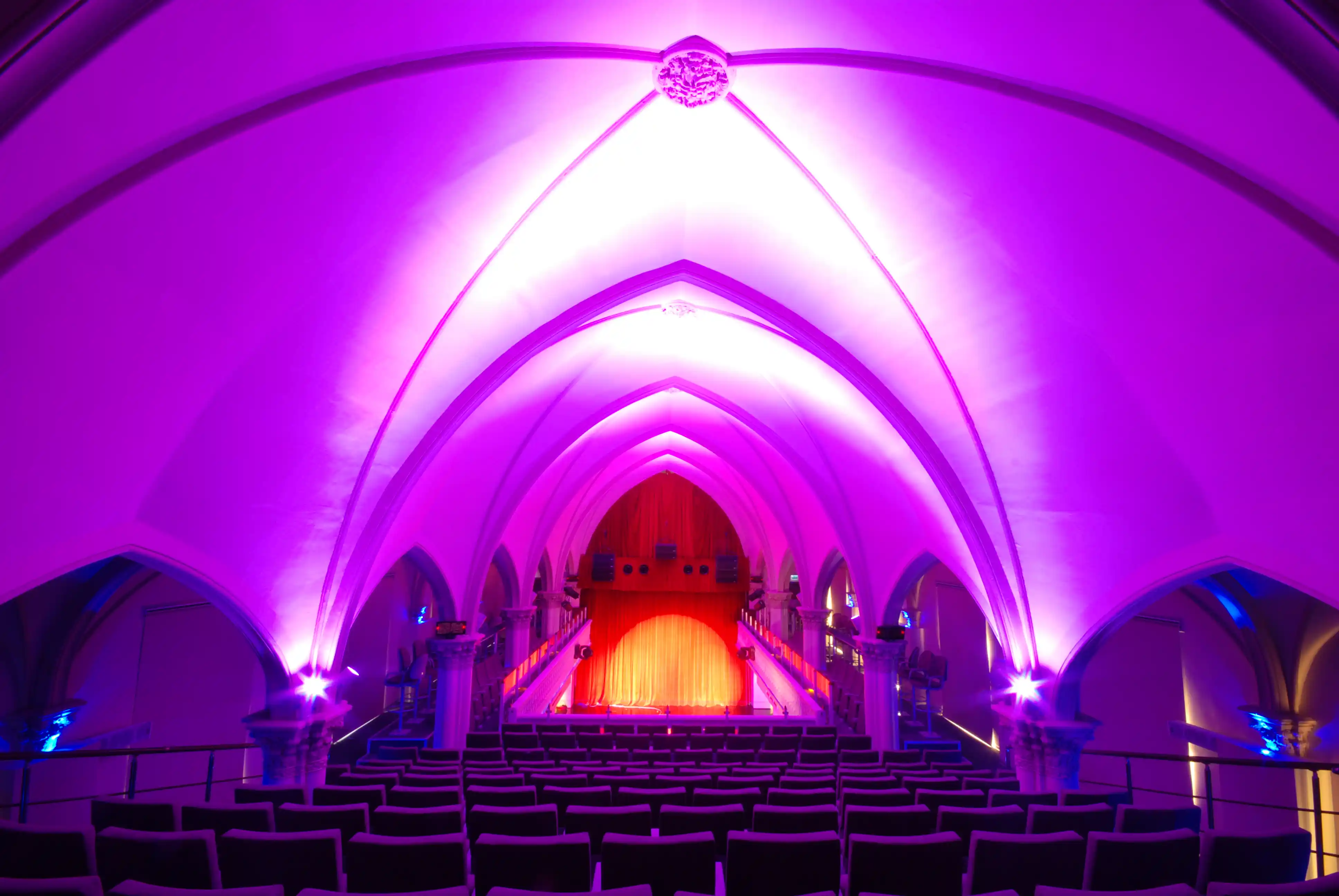 Interior of a Gothic-style auditorium with vaulted ceilings lit in purple and a red curtain stage at the front.