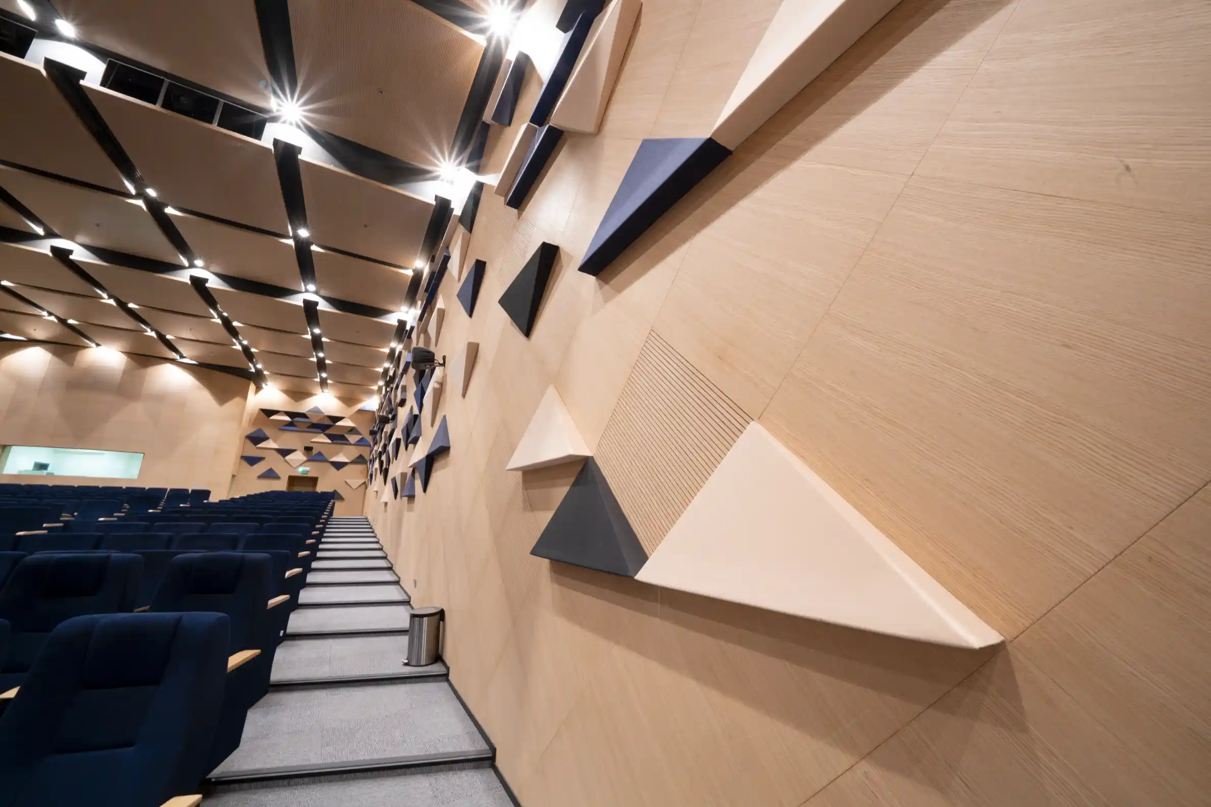 Modern auditorium with blue seats, wooden walls featuring triangular acoustic panels, and ceiling with recessed lighting.
