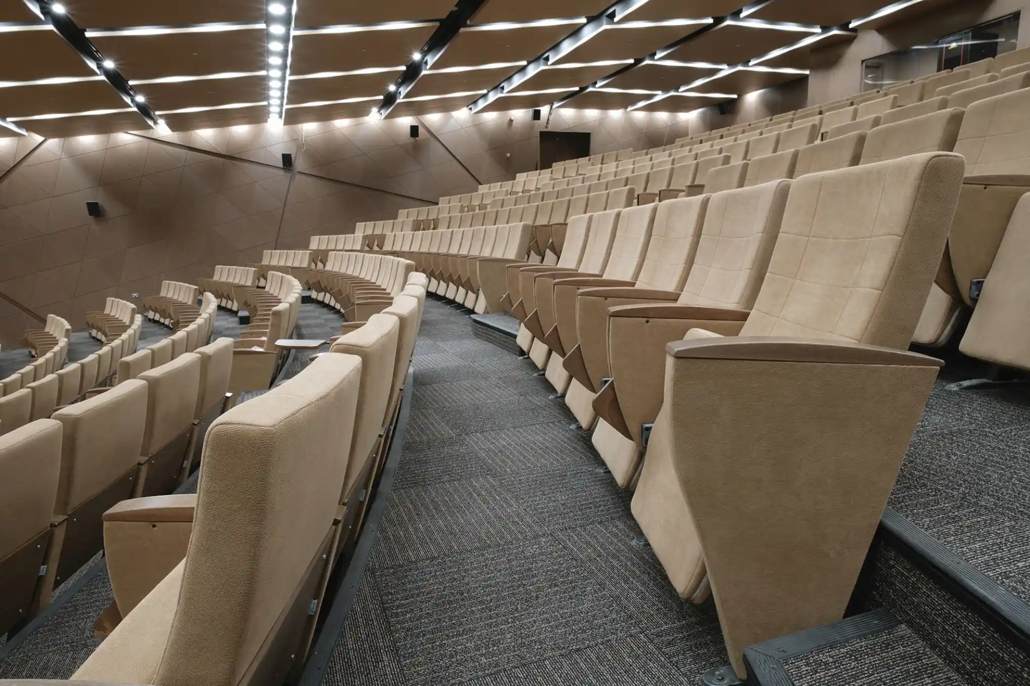 Modern auditorium with multiple rows of beige upholstered seats and angled ceiling lights.