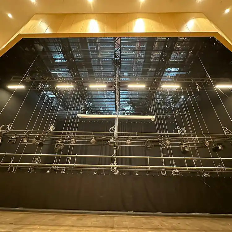 Stage lighting rig with multiple hanging spotlights and cables against a black backdrop and wooden ceiling.