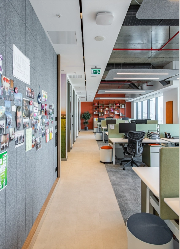 Modern open-plan office with workstations, ergonomic chairs, a bulletin board with photos on the left wall, and a large window on the right letting in natural light.
