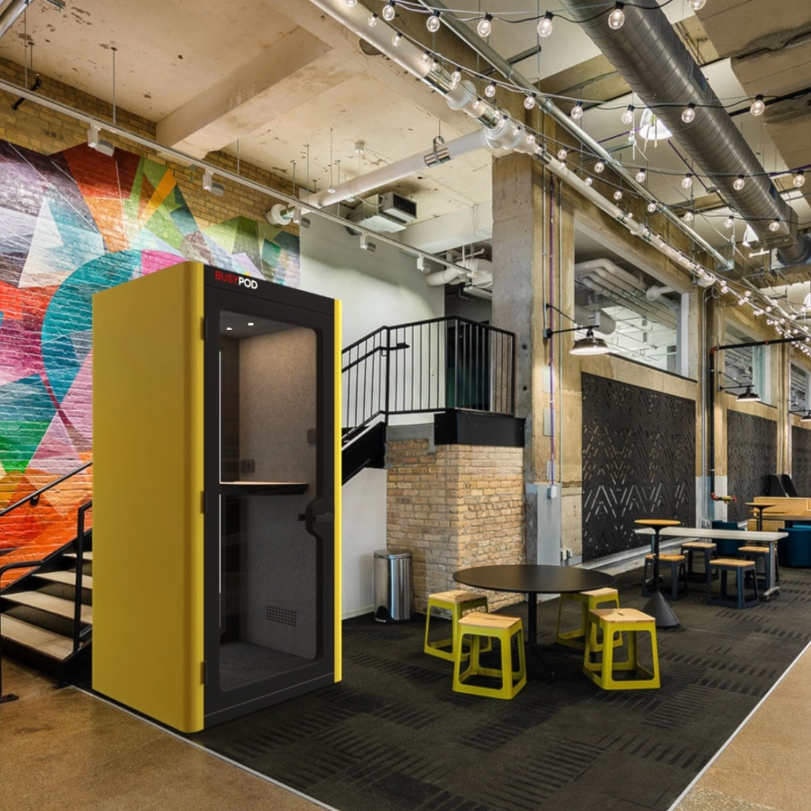 Modern office space with a yellow BusyPod acoustic phone booth, colourful wall mural, round table with yellow stools, and string lights overhead.