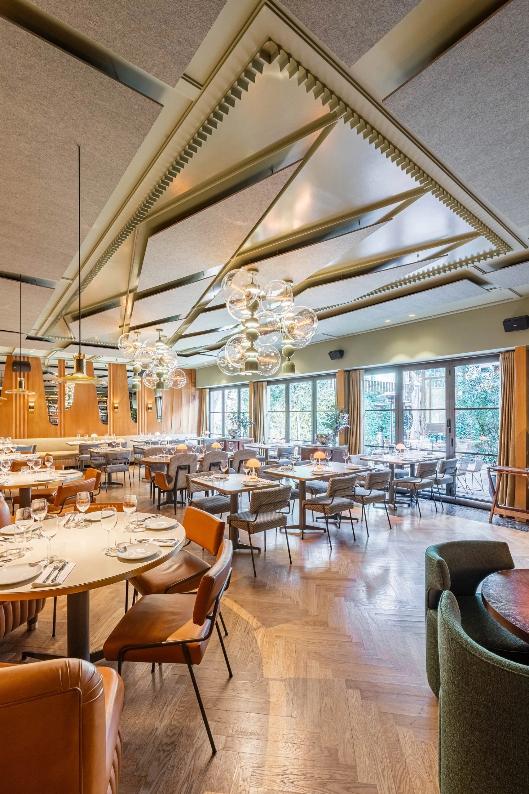 Bright, modern restaurant interior with wooden floors, round and square tables set with glasses and plates, leather and fabric chairs, large windows, and decorative ceiling lighting.