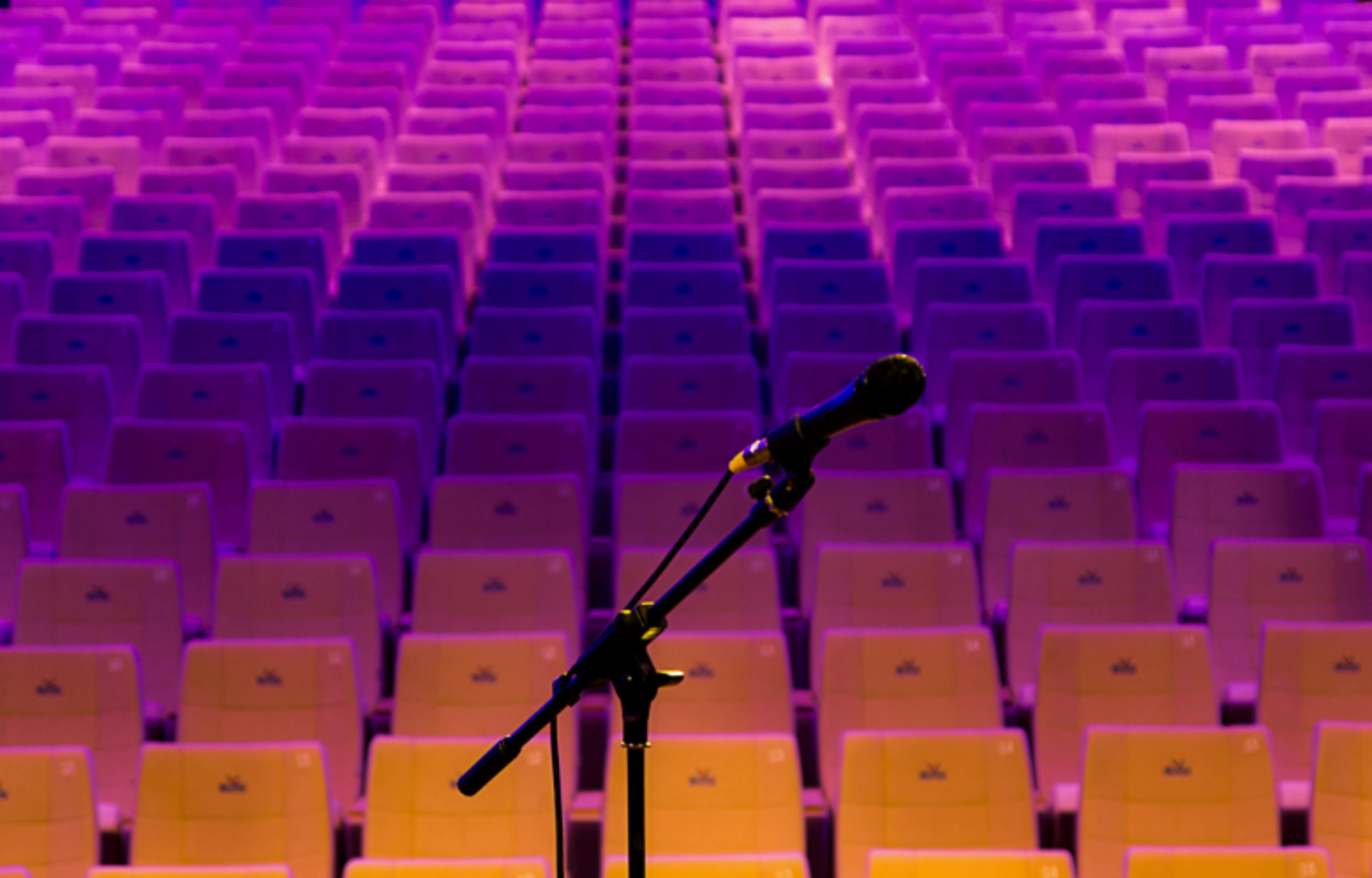 Microphone on a stand in front of empty theatre seats illuminated with purple and orange lighting.