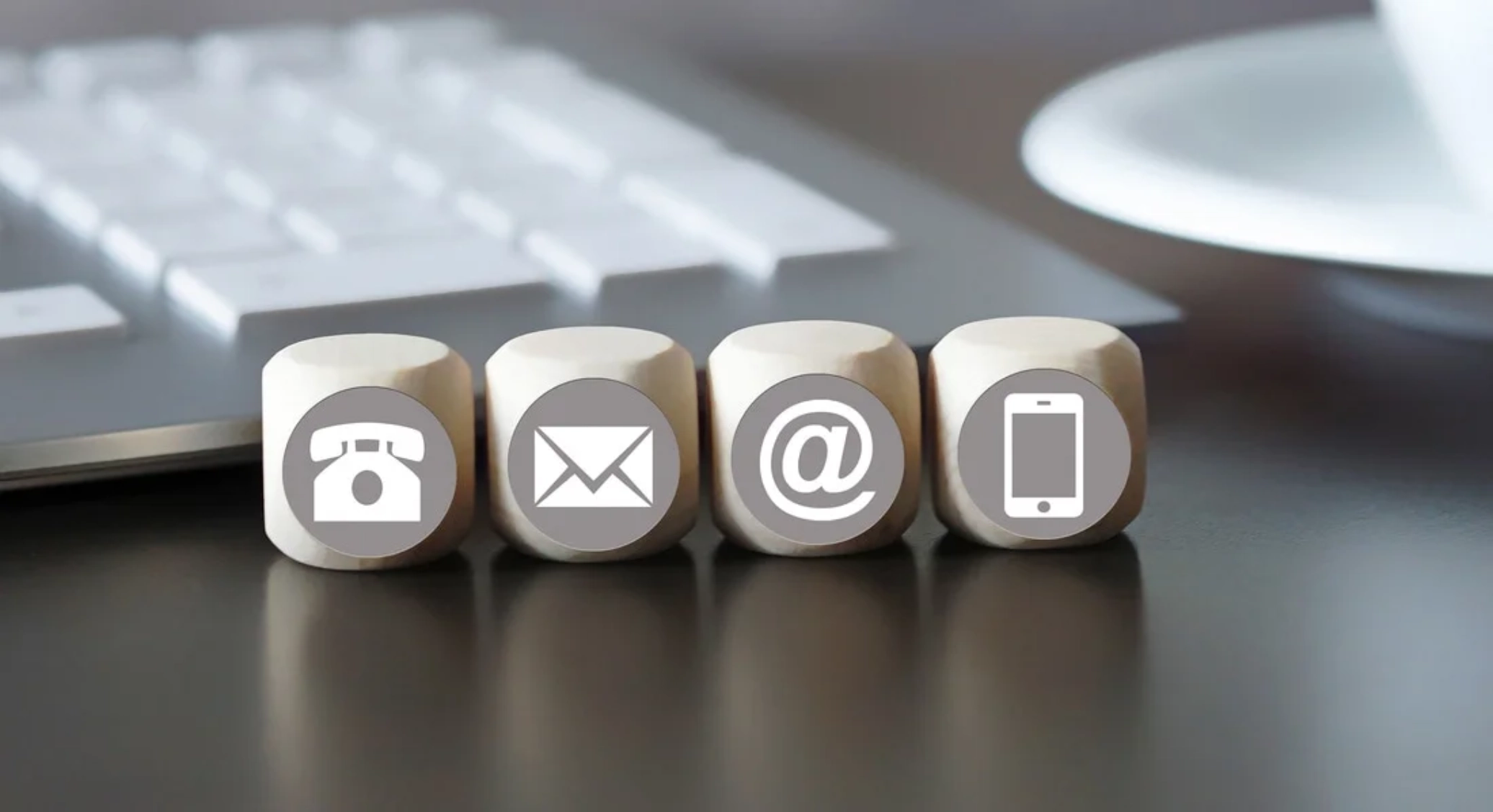 Four wooden blocks with icons for telephone, email, at symbol, and mobile phone arranged in front of a computer keyboard.