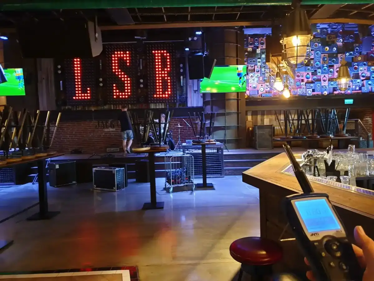 Empty bar with tables and chairs stacked, illuminated LSB sign on the wall, TV screens, and a sound measurement device held in the foreground.