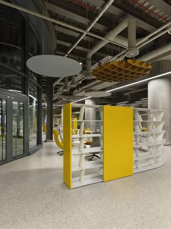 Modern open office space with yellow and white shelving units, grey circular and brown grid-patterned acoustic ceiling panels, and exposed ceiling pipes.