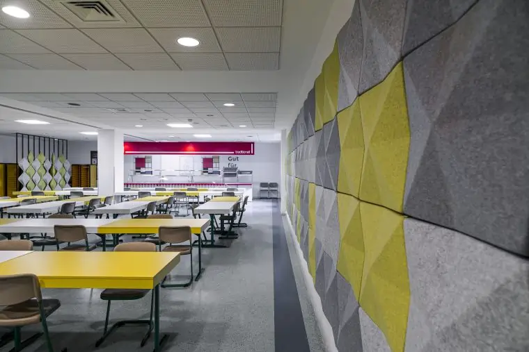 Modern cafeteria with yellow tables, beige chairs, and a grey and yellow geometric acoustic wall panel on the right.