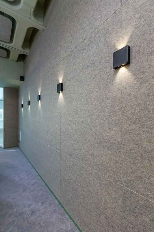 Long gray felt acoustic wall with six evenly spaced black wall-mounted lights casting upward and downward beams.
