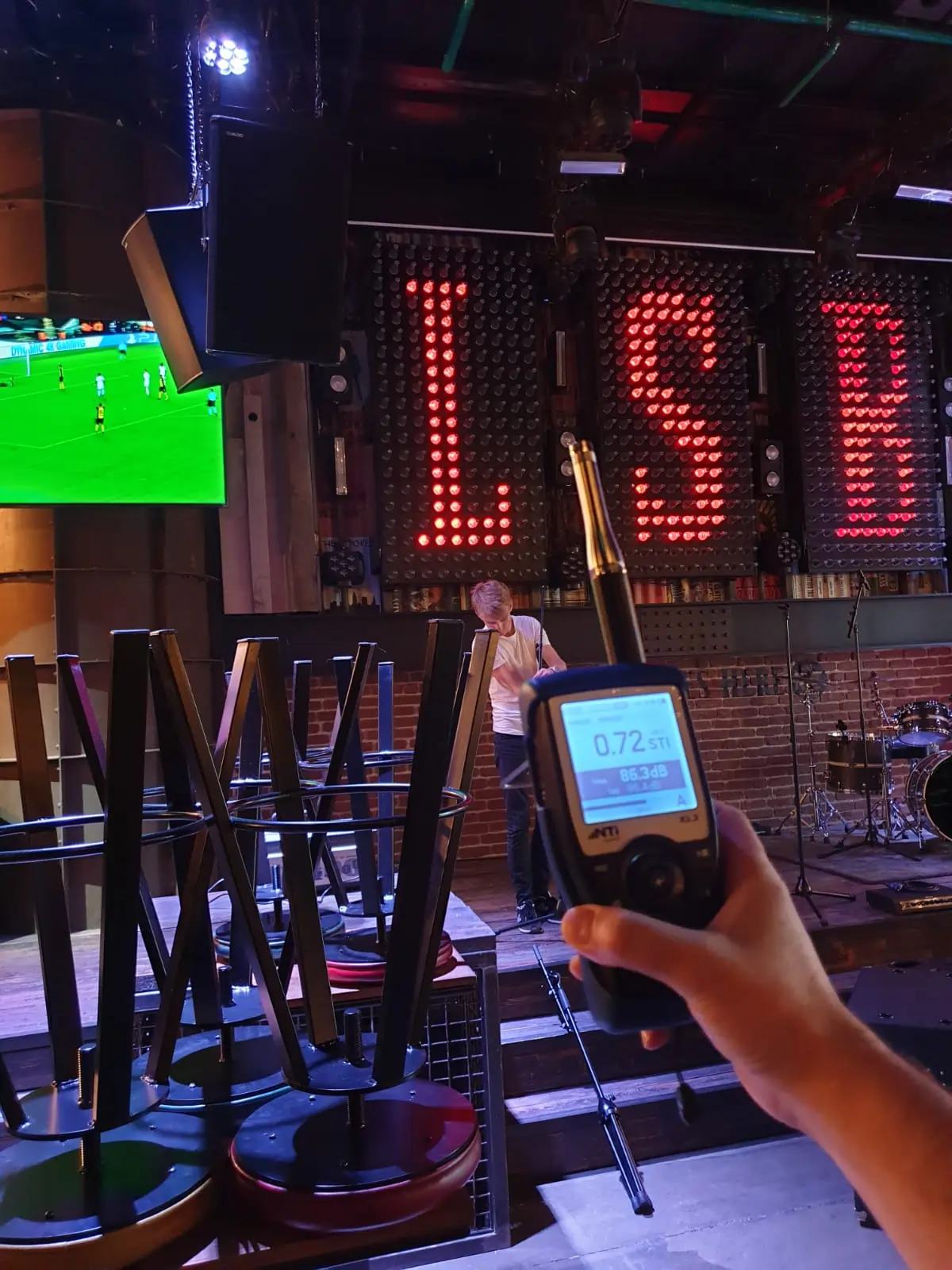 Hand holding a sound level meter in a dimly lit music venue with stacked chairs, stage, drum set, and a large illuminated 'LSD' sign in the background.