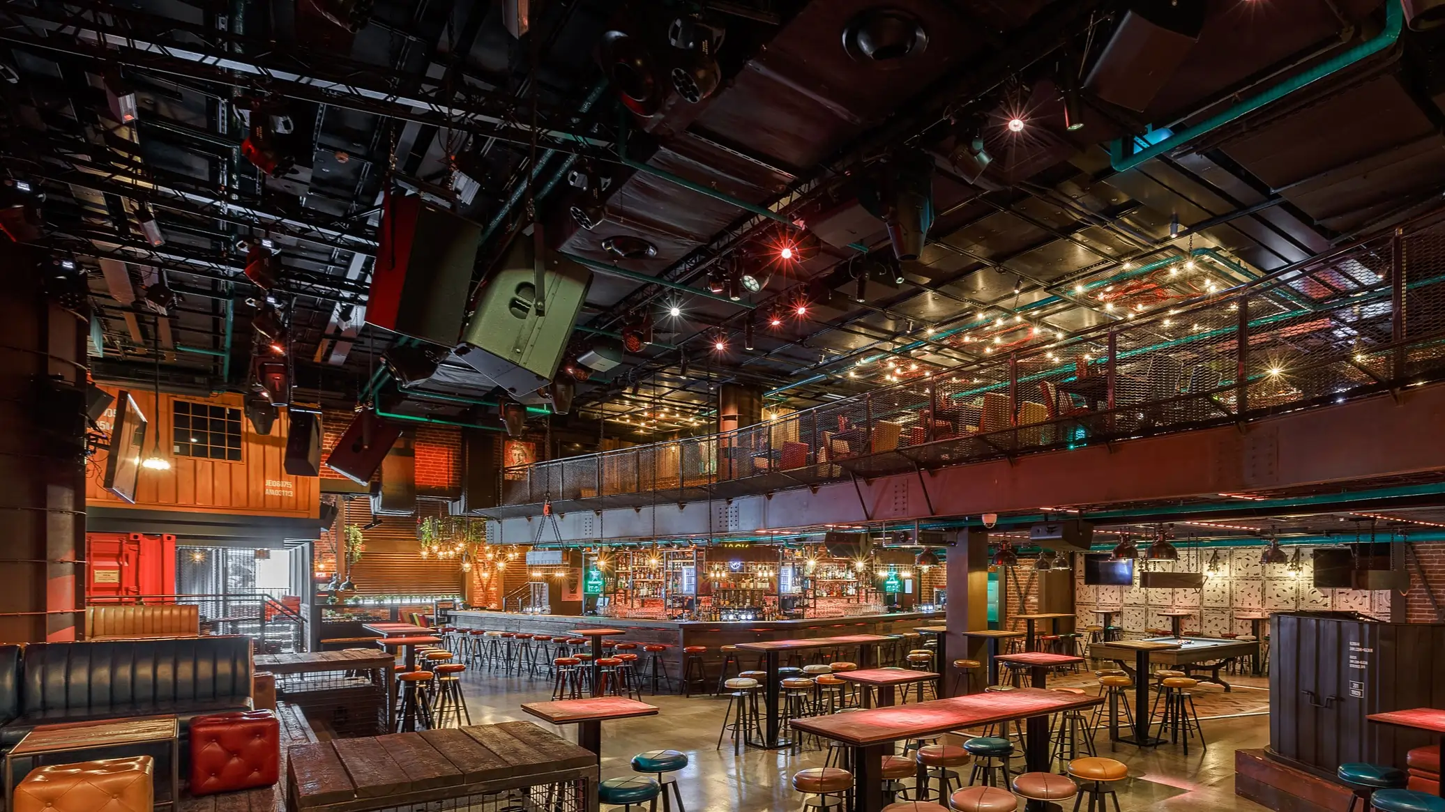 Spacious modern bar interior with wooden tables, colourful stools, lit hanging bulbs, and upper balcony seating area.