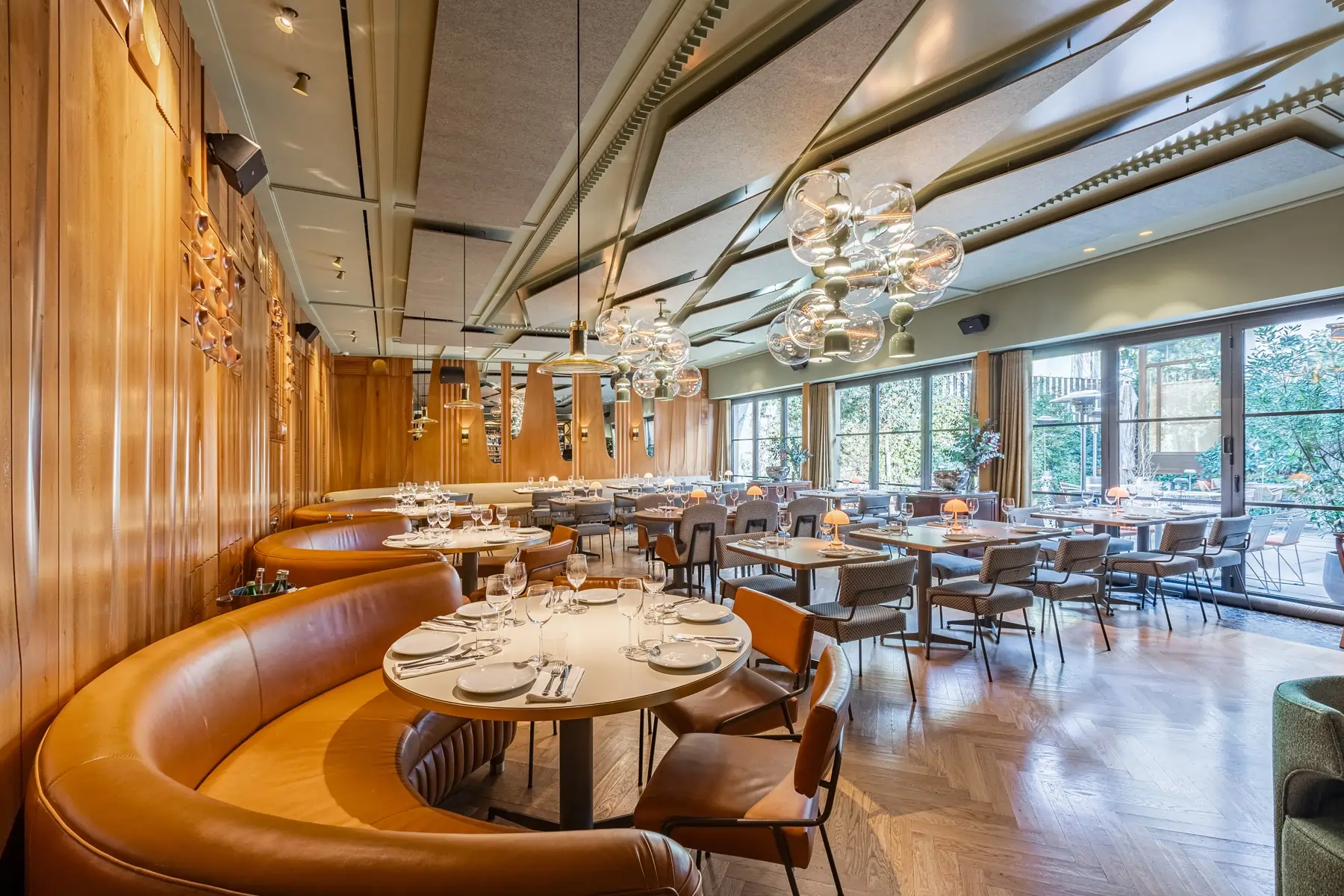 Modern restaurant interior with curved leather banquettes, round tables set with plates and glasses, pendant lighting, and large windows overlooking a garden patio.