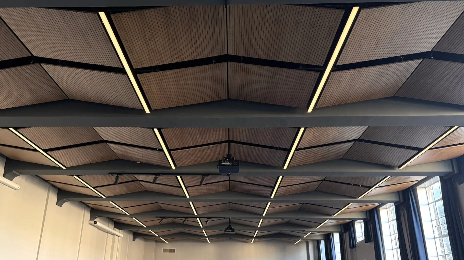 Indoor ceiling with wooden acoustic panels, linear lights, and large windows with dark curtains on the right.