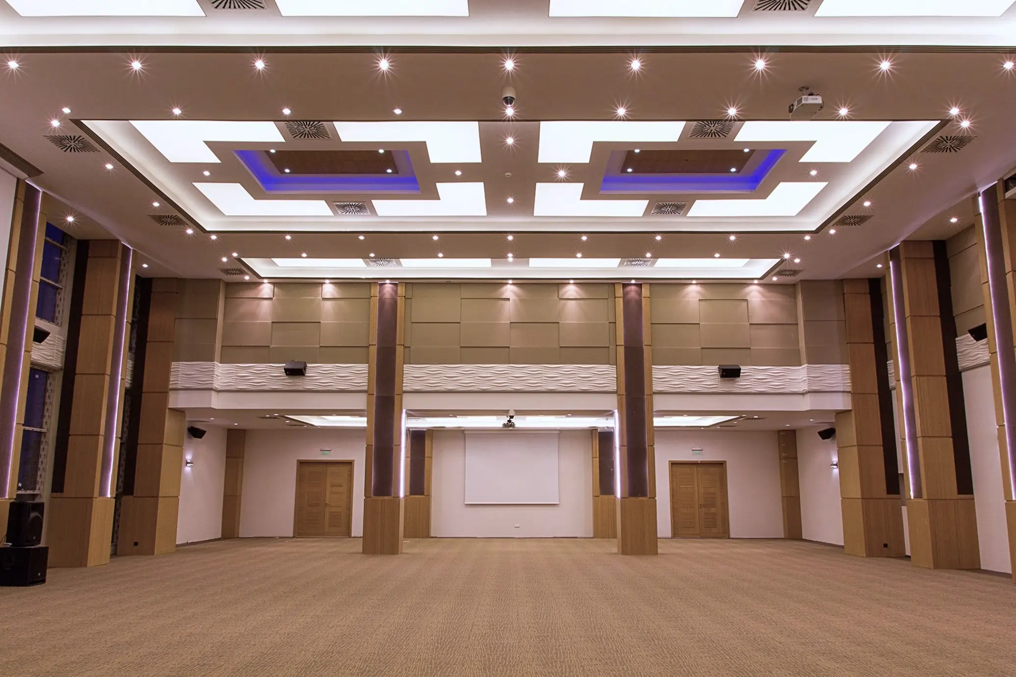 Spacious modern conference hall with beige carpeting, wooden columns, recessed ceiling lights, and a projection screen at the front.