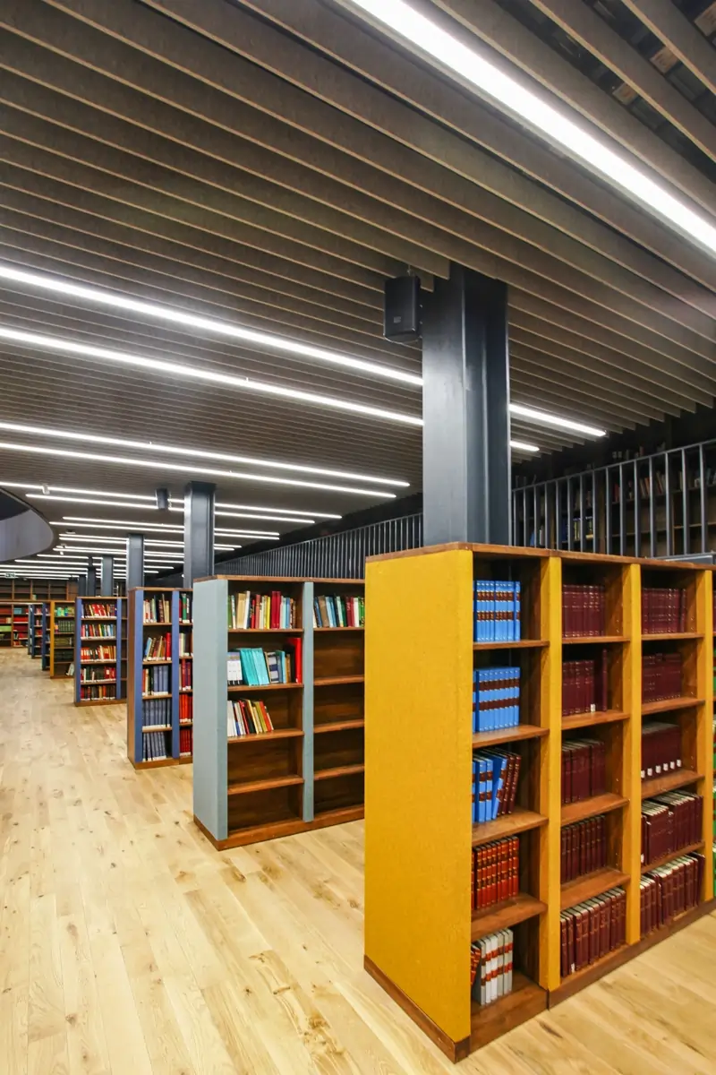 Rows of wooden bookshelves filled with books under a modern ceiling with linear lights in a spacious library.