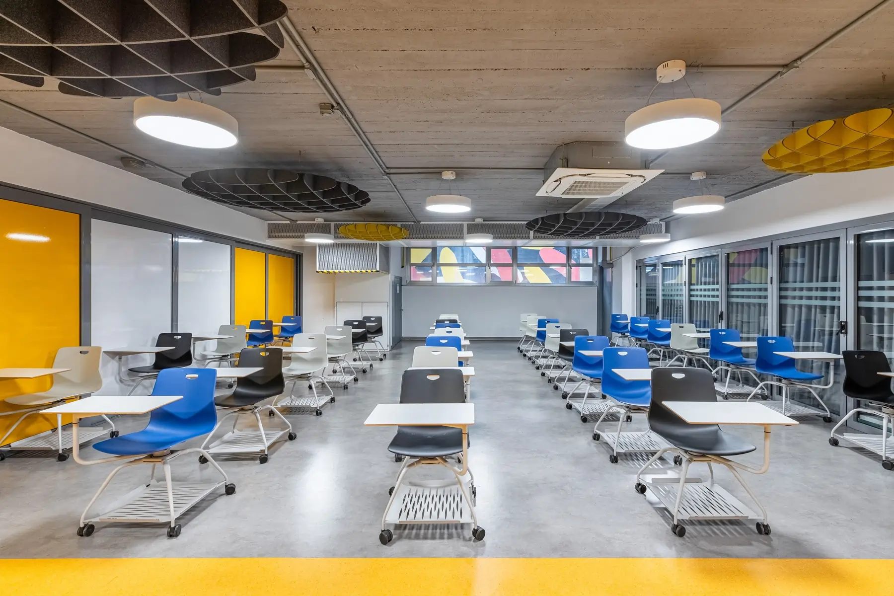 Modern empty classroom with rows of colourful blue, black, and white chairs with attached desks on wheels under circular ceiling lights.