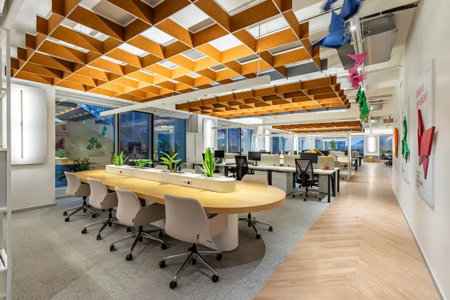 Modern open-plan office with wooden ceiling panels, long curved desks with plants, and ergonomic chairs.