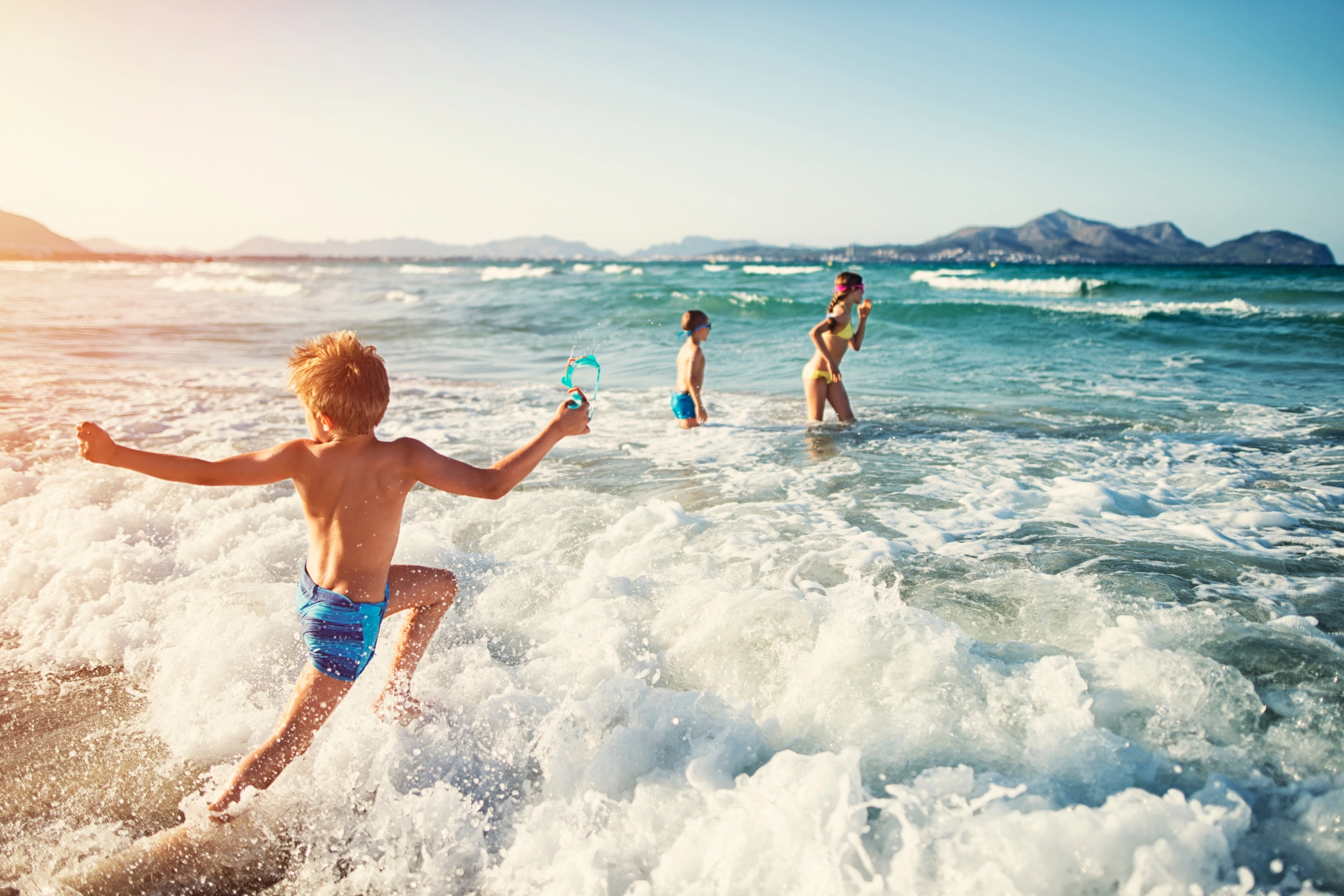 Kinder spielen im Meerwasser am Strand bei sonnigem Wetter mit Bergen im Hintergrund.