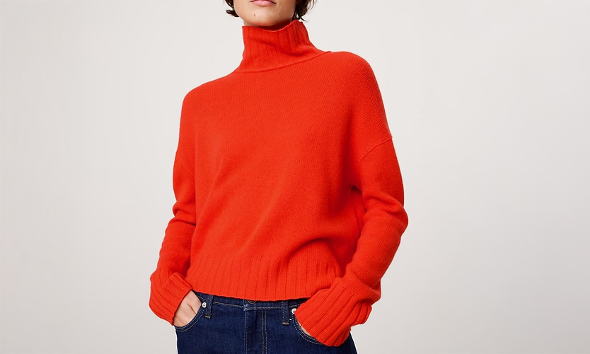 Person wearing a bright orange turtleneck sweater and dark blue jeans with hands in pockets against a plain light background.