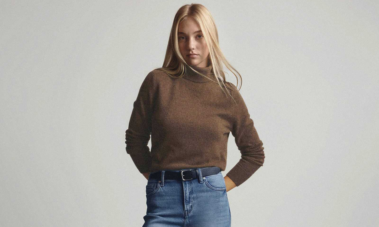 Blonde woman wearing a brown turtleneck sweater tucked into high-waisted blue jeans with a black belt, standing against a plain light background.