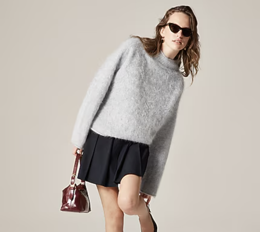 Woman wearing a light gray fuzzy sweater, black pleated skirt, sunglasses, and holding a small maroon handbag.