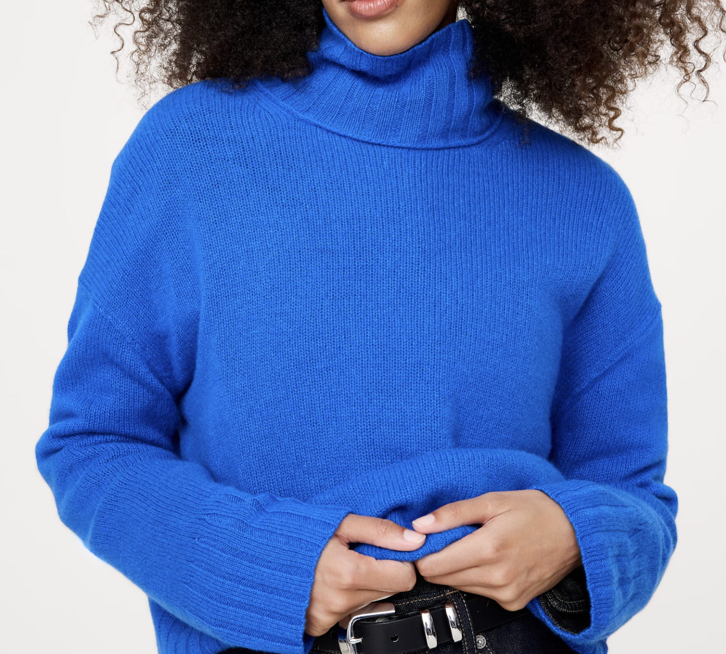 Person wearing a bright blue turtleneck sweater holding the hem near the waist over dark jeans with a black belt.