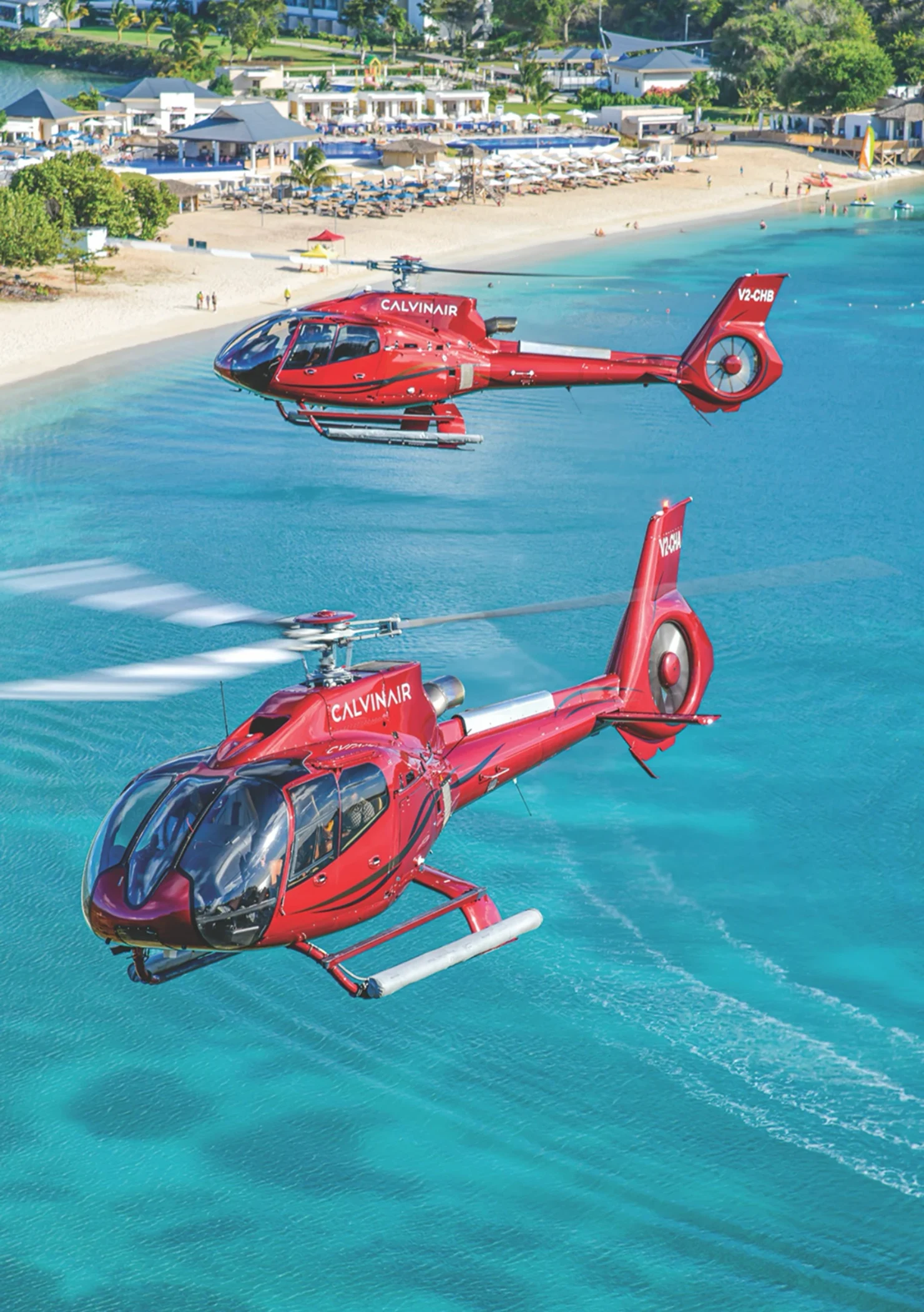 Two red helicopters flying over turquoise waters near a tropical beach