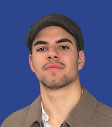 Young man with short beard wearing a brown flat cap and brown jacket against a blue background.