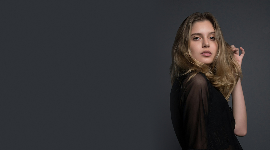 Young woman with long blonde hair wearing a black sheer top posing against a dark gray background.