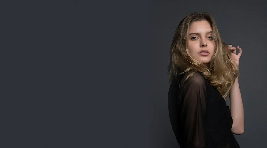 Young woman with long blonde hair wearing a black sheer top posing against a dark gray background.