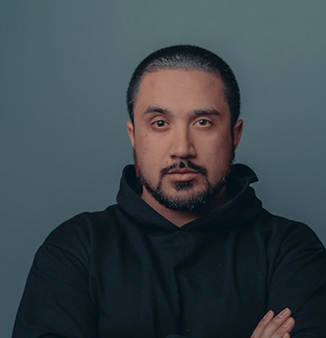 Man with short hair and beard wearing a black hoodie, standing against a plain gray background with arms crossed.