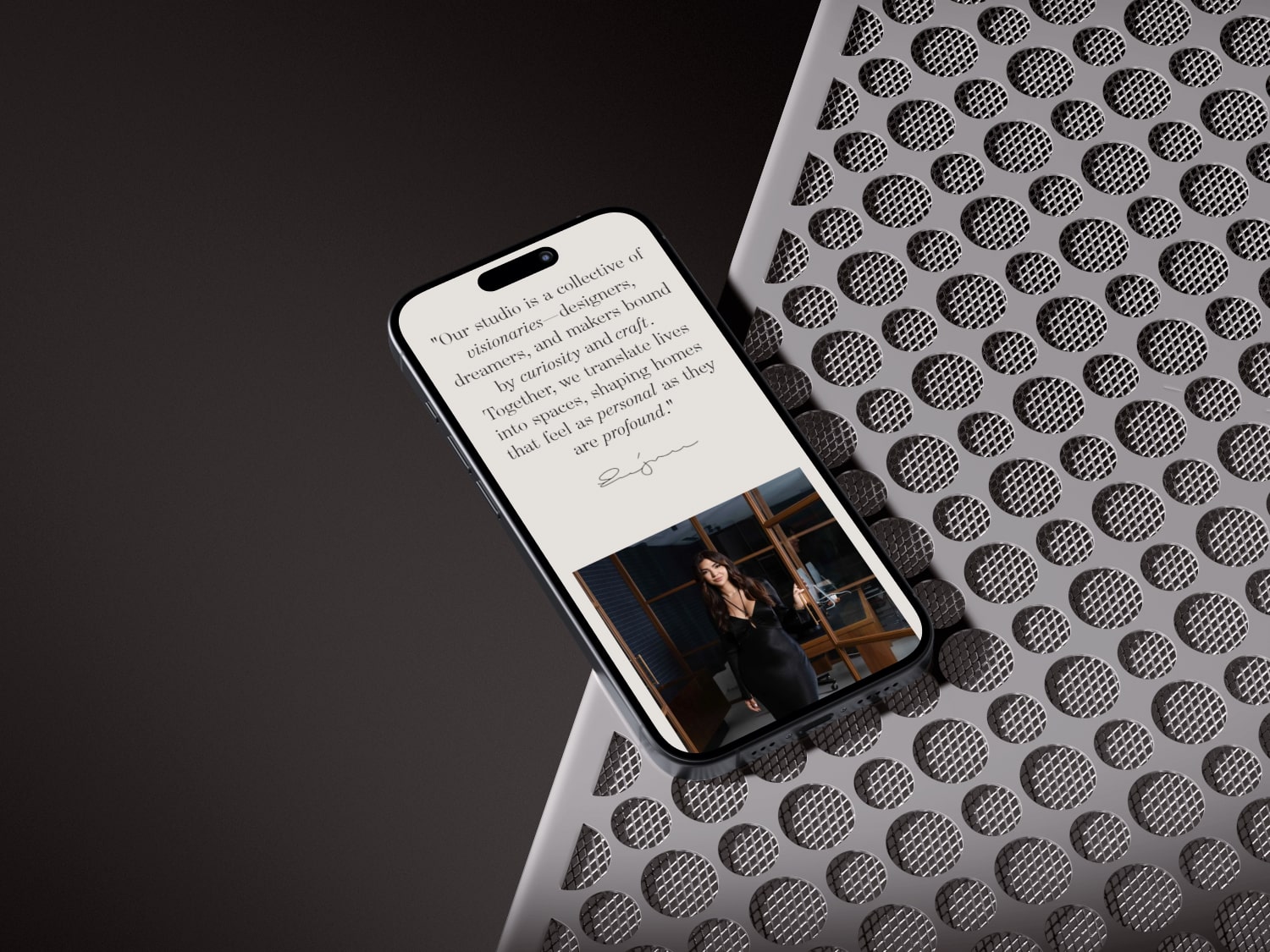 Smartphone displaying a quote about a design studio and a photo of a woman in a black dress standing by windows, placed on a metal surface with circular perforations.