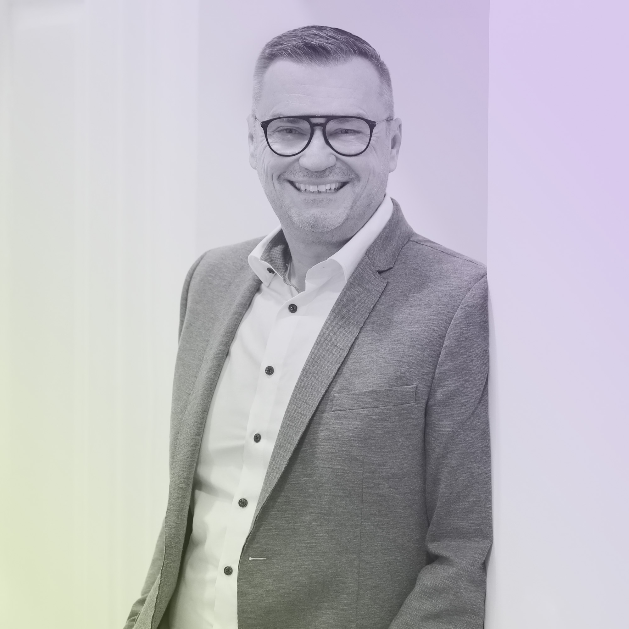 Ein älterer Mann mit kurzen Haaren und Brille lächelt in grauem Anzug und weißem Hemd, lehnt sich an eine weiße Wand.