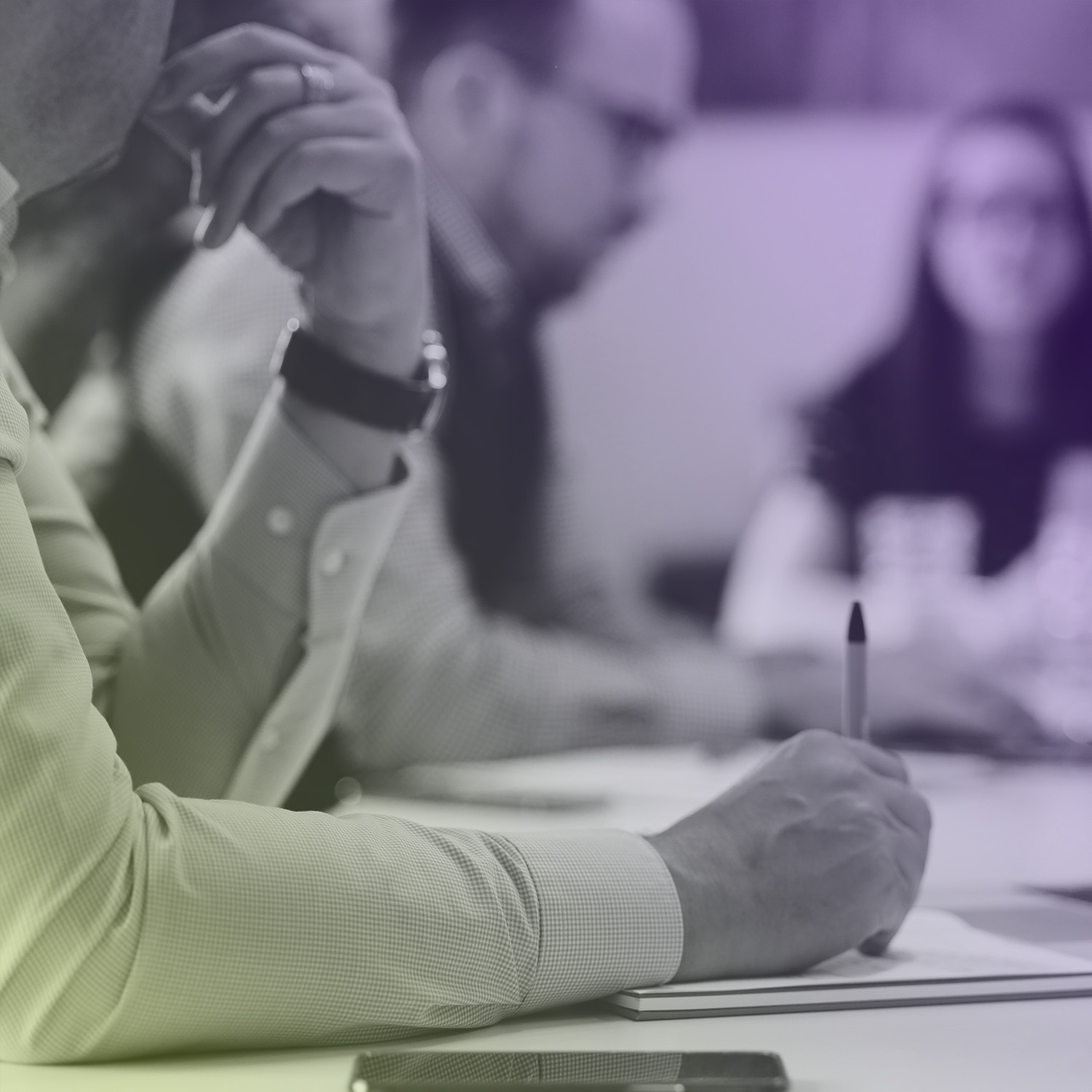 Person schreibt mit Kugelschreiber auf einem Notizblock während eines Meetings mit zwei weiteren Personen am Tisch.