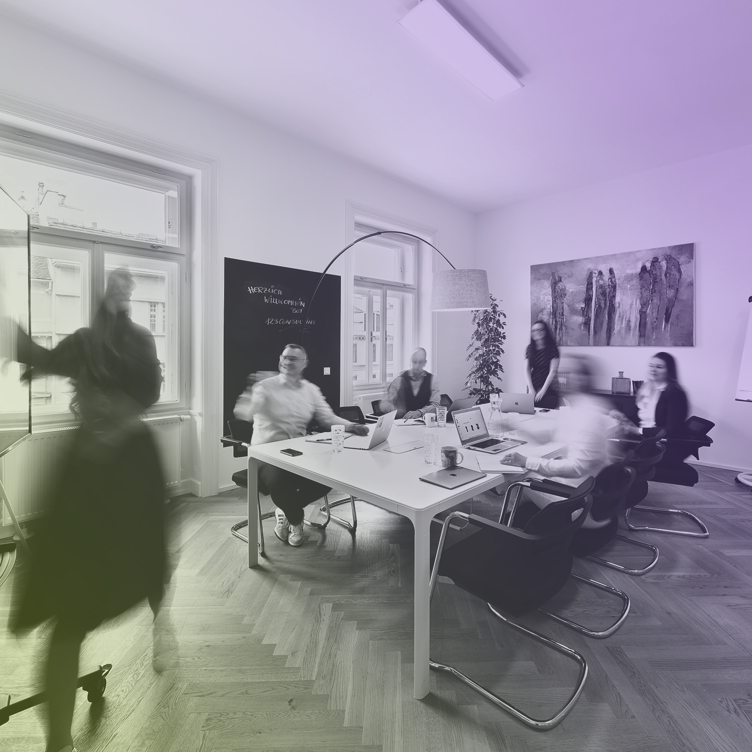 Bürobesprechung mit sechs Personen an einem Tisch mit Laptops in einem modernen Raum.