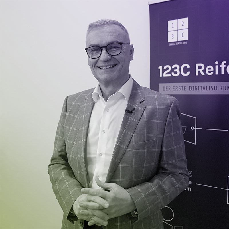 Ein älterer Mann mit kurzen Haaren und Brille lächelt in grauem Anzug und weißem Hemd, lehnt sich an eine weiße Wand.