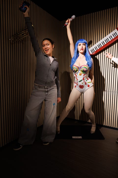Une femme pose à côté d'une statue de cire d'une chanteuse avec une perruque bleue et un costume coloré, toutes deux levant un bras avec un micro.