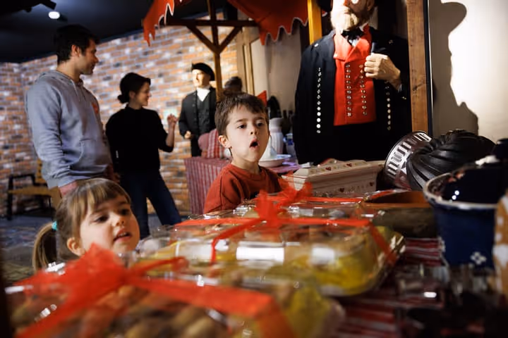 Deux enfants regardant des pâtisseries emballées avec des rubans rouges sur une table, avec deux adultes discutant en arrière-plan dans un environnement de marché ou boutique.