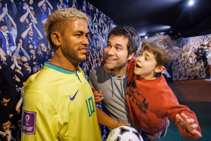Un homme et un enfant regardant avec admiration une statue de cire de Neymar en maillot jaune du Brésil, avec un ballon de football.