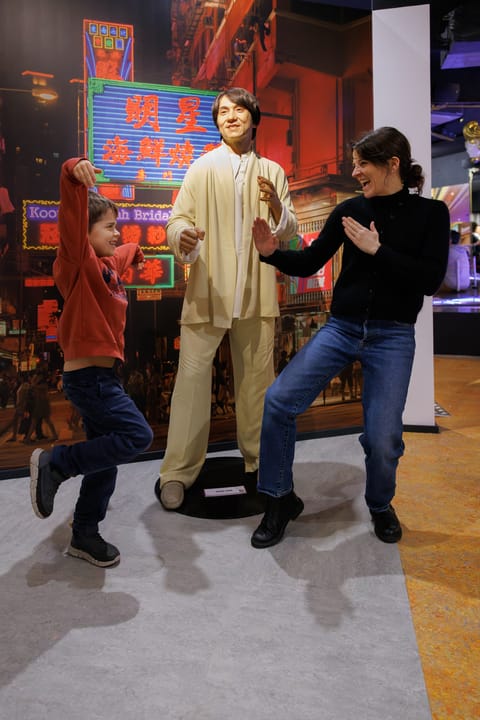 Un enfant et une femme imitent la posture d'un homme en statue habillé en tenue beige devant un fond urbain lumineux avec des enseignes chinoises.