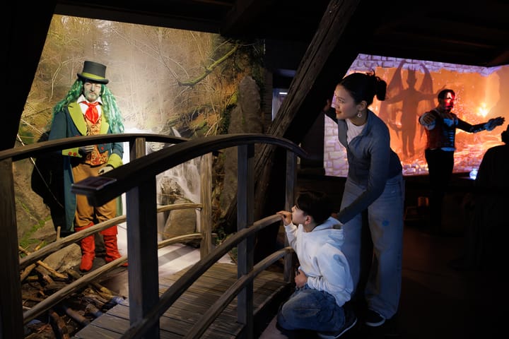 Enfant en sweat à capuche blanche avec une femme penchée vers lui, observant un personnage en costume coloré et haut-de-forme sur un pont dans une exposition muséale.